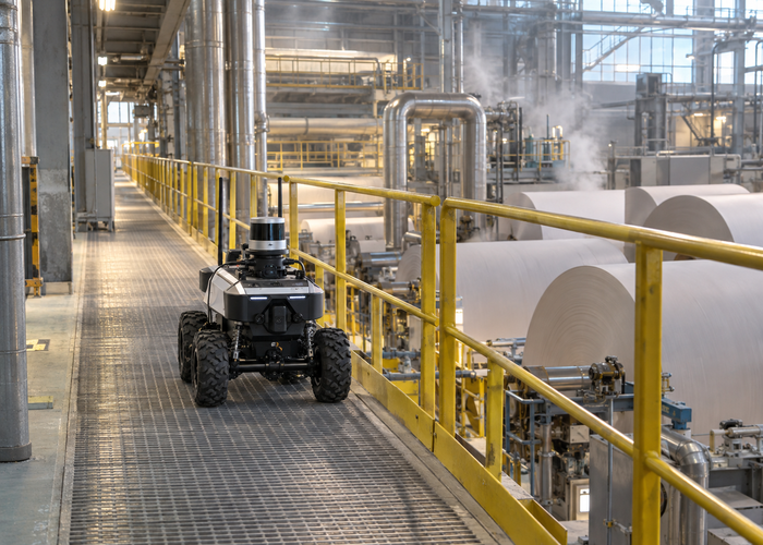 rugged mobile inspection robot with a physical roof-mounted LiDAR sensor traveling on a guarded paper-mill walkway beside large paper rolls and steam pipes in a realistic LiDAR application scene
